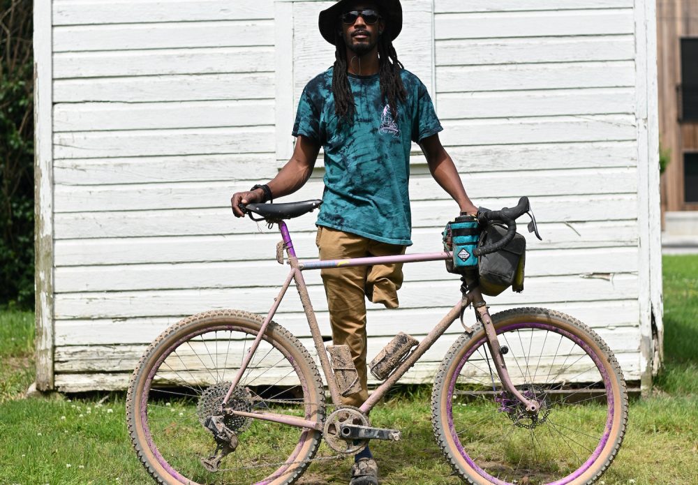 Leo Rogers, a black man with one leg, stands suavely with his bike against a white shed