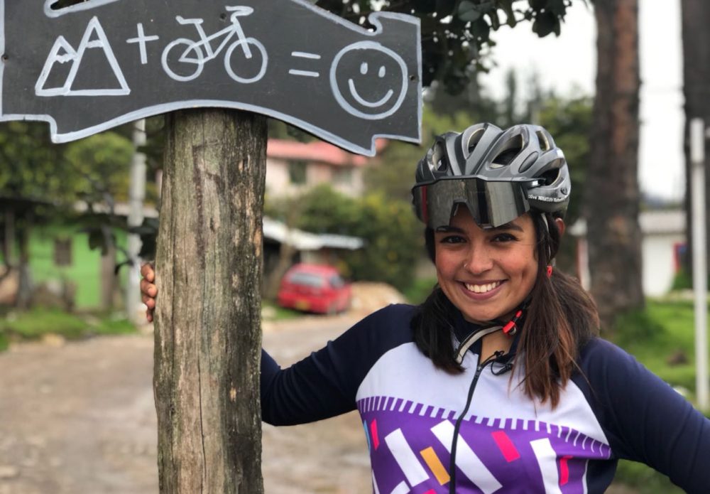 Latin American female in cycling outfit next to a sign that shows a mountain, bicycle and smiley face