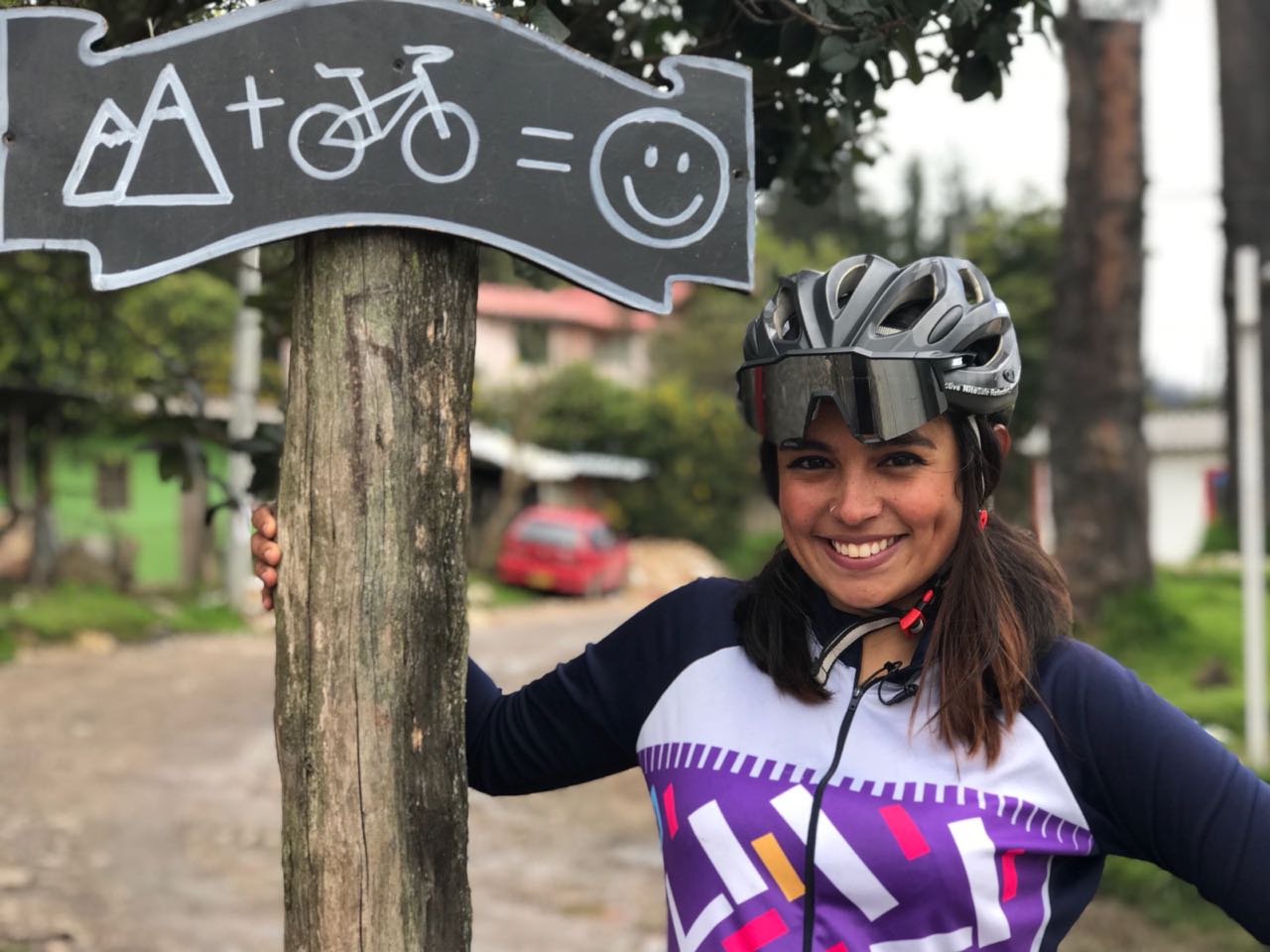 Latin American female in cycling outfit next to a sign that shows a mountain, bicycle and smiley face