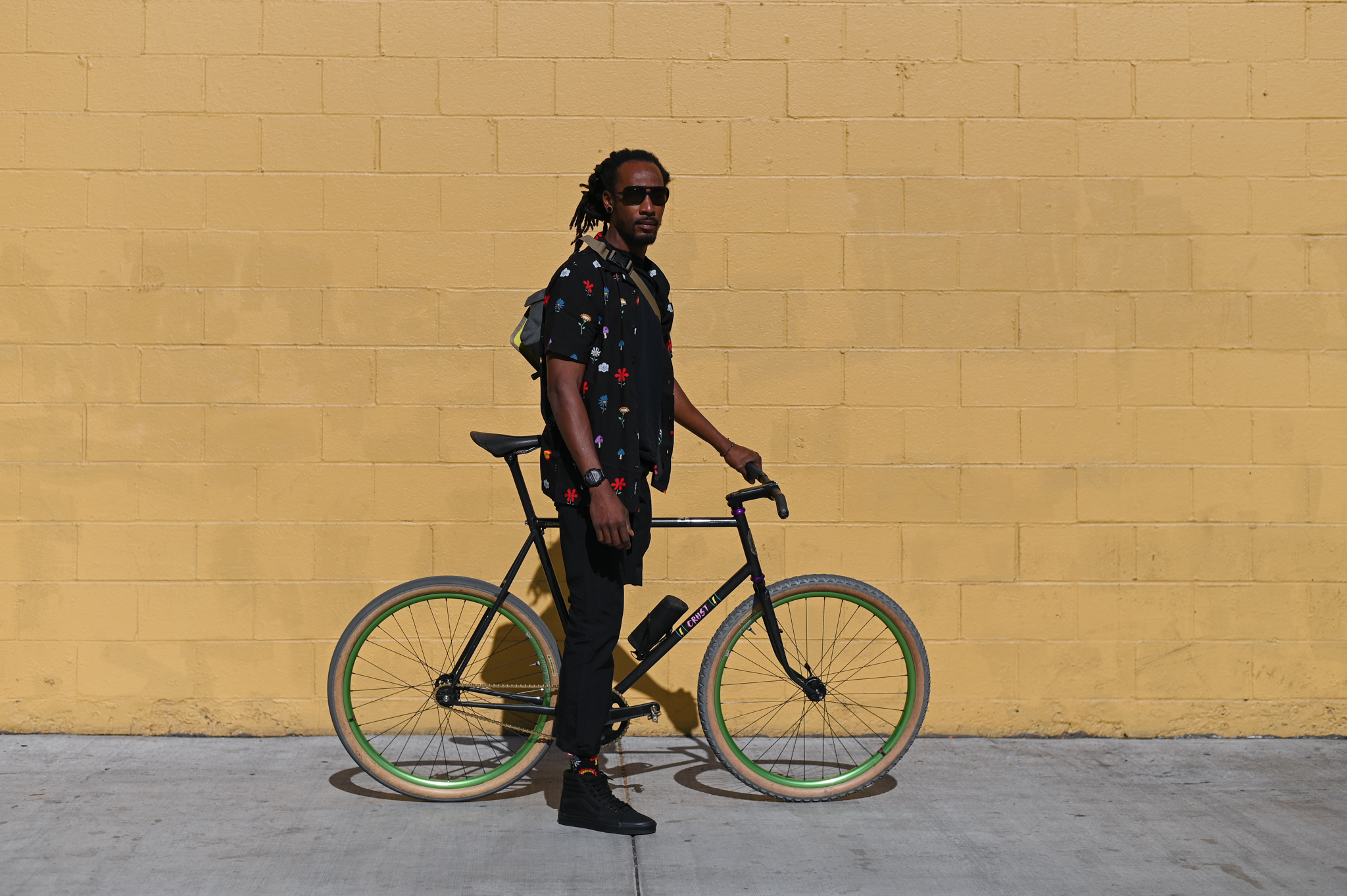 Leo, a black man with dreadlocks and one leg, is wearing slick black clothes and standing with his bike against a beige brick wall